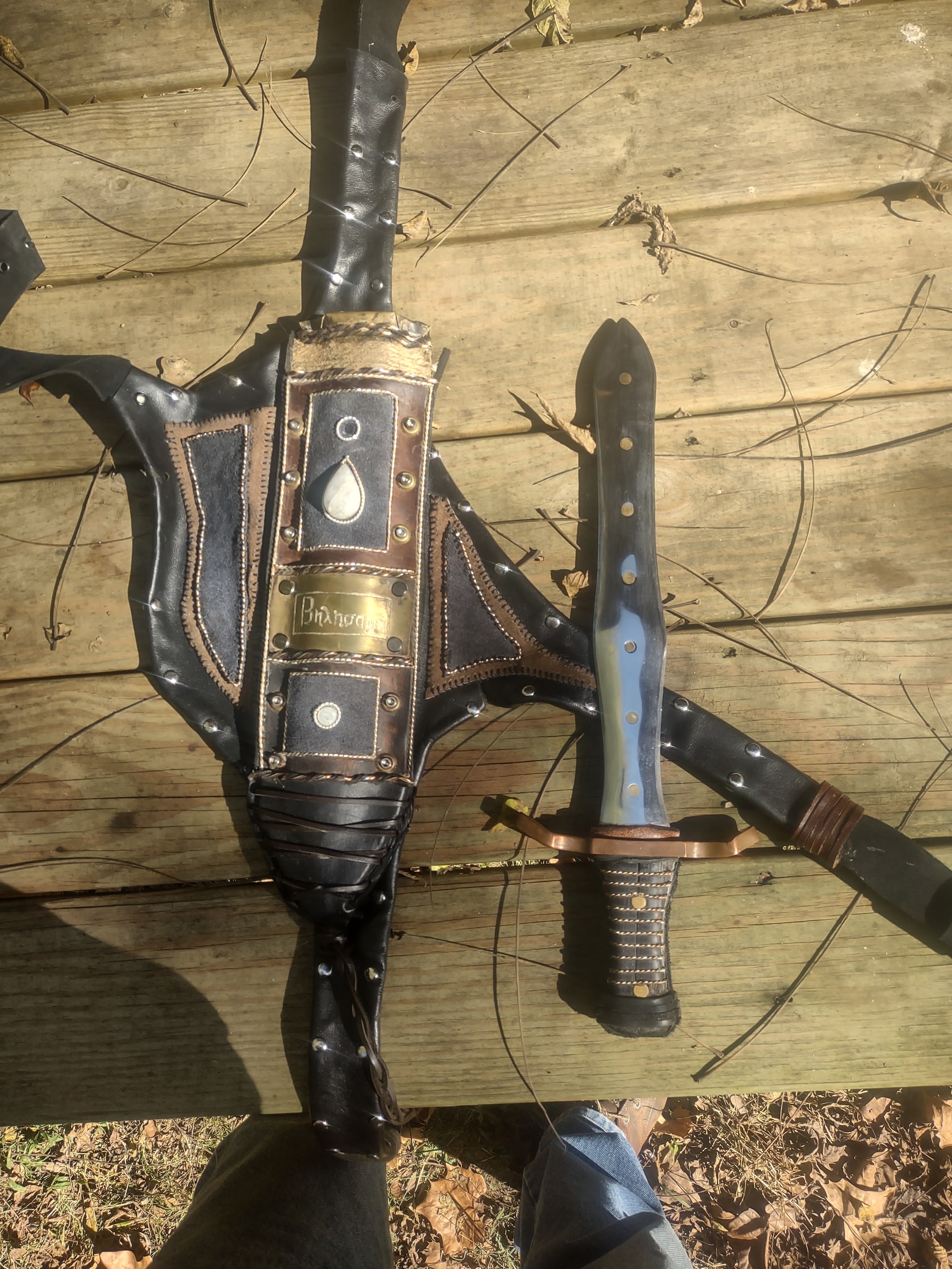 Ornate leather sheath with brass plate alongside blue-handled knife on wood deck