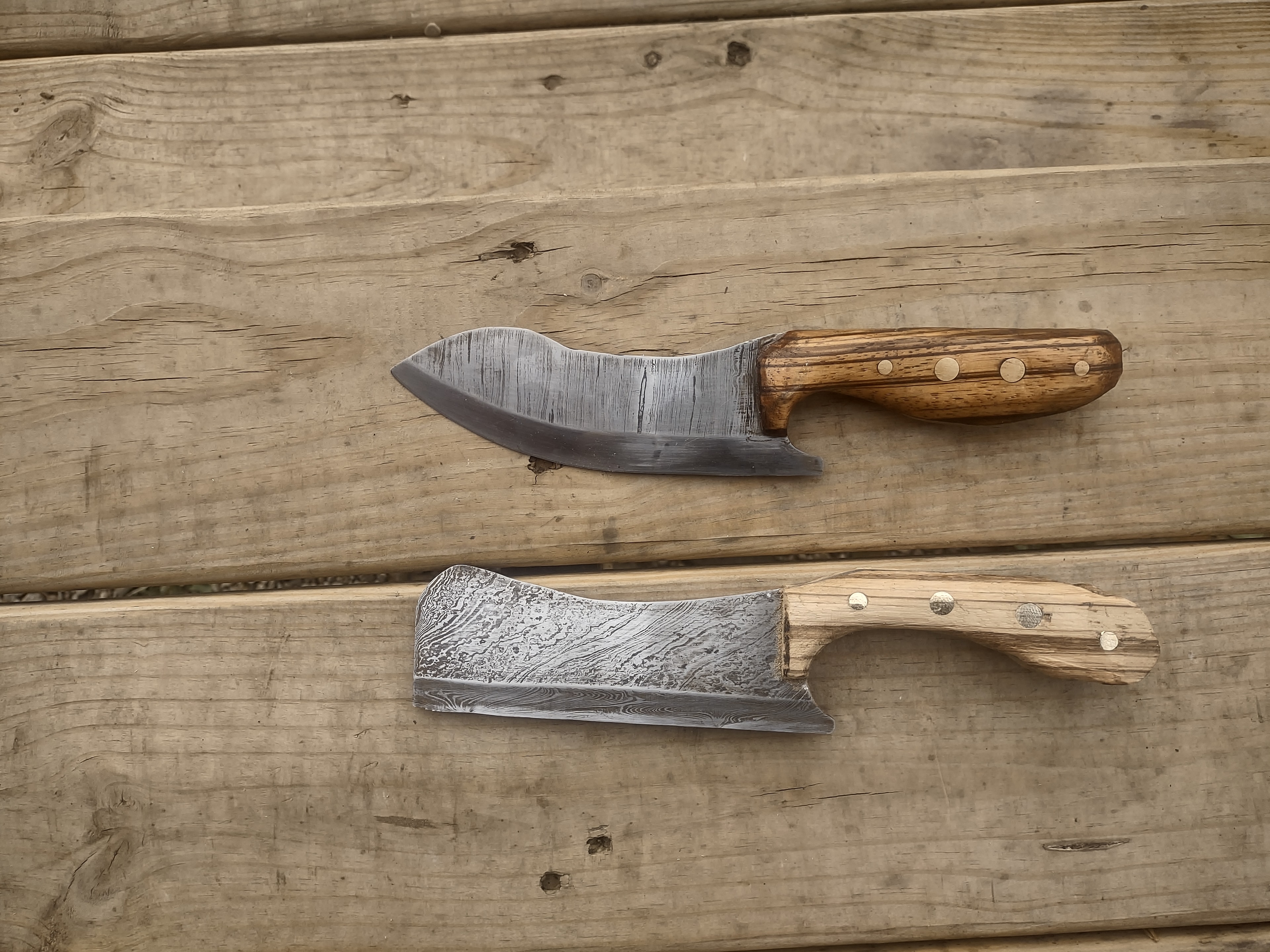 Two handcrafted knives on wood deck — curved blade with oak handle and Damascus cleaver with ash handle